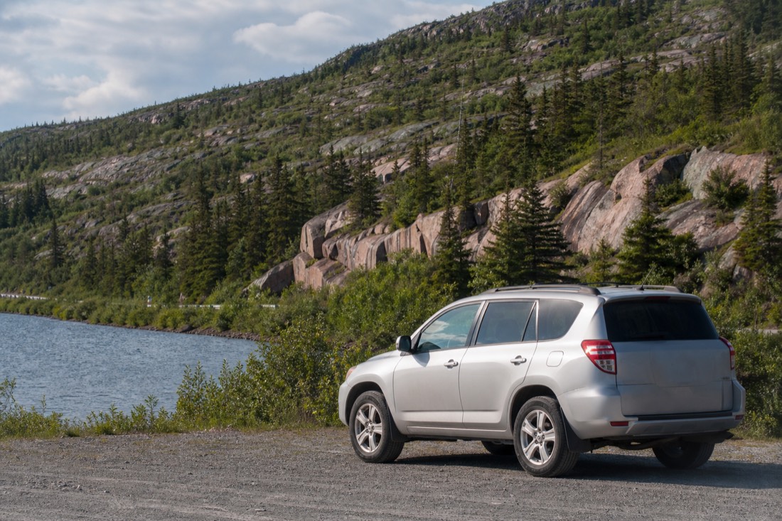 Rental SUV parked at a gravel pullout beside a lake on Highway 4 east of Yellowknife with spruce and granite behind