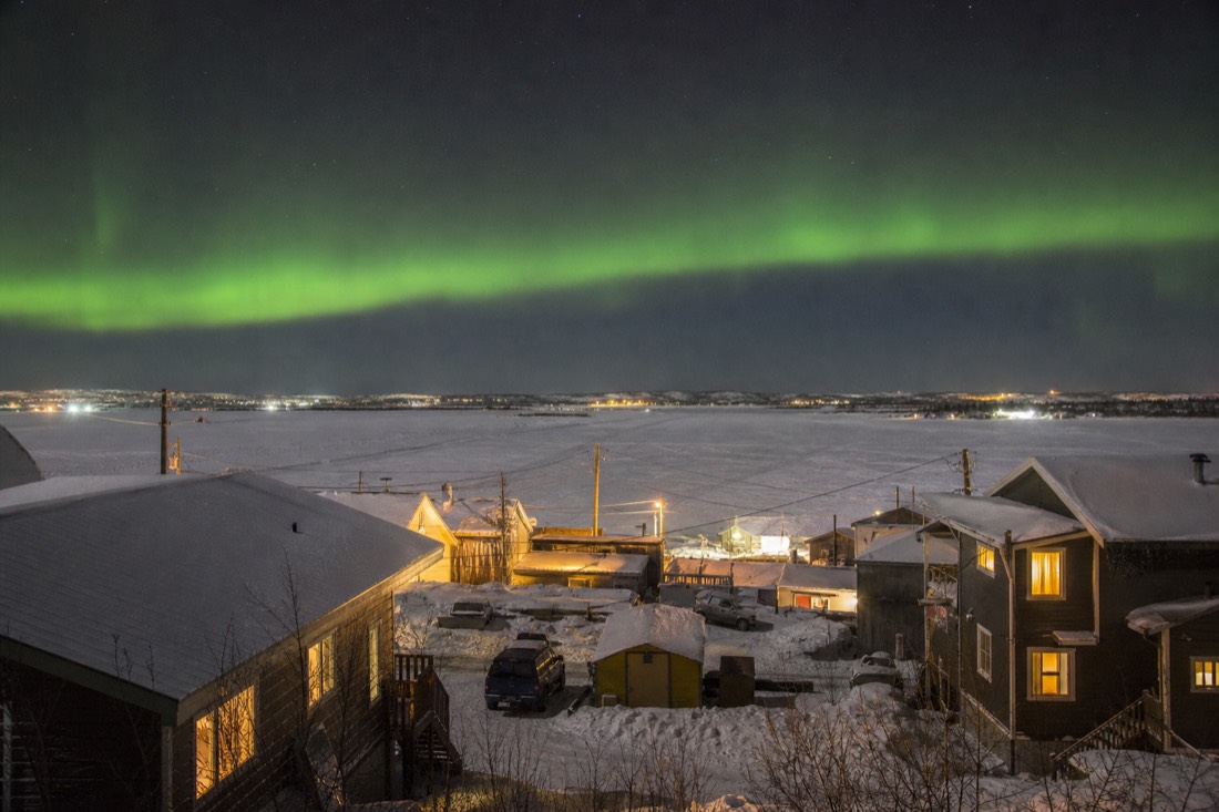 Winter afternoon in Old Town Yellowknife with warm window lights and the frozen bay glowing beyond the shacks