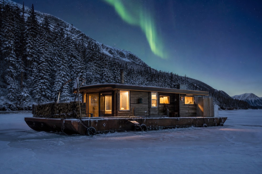 Houseboat frozen into the bay ice in deep winter with firewood stacked on deck and lit windows at dusk