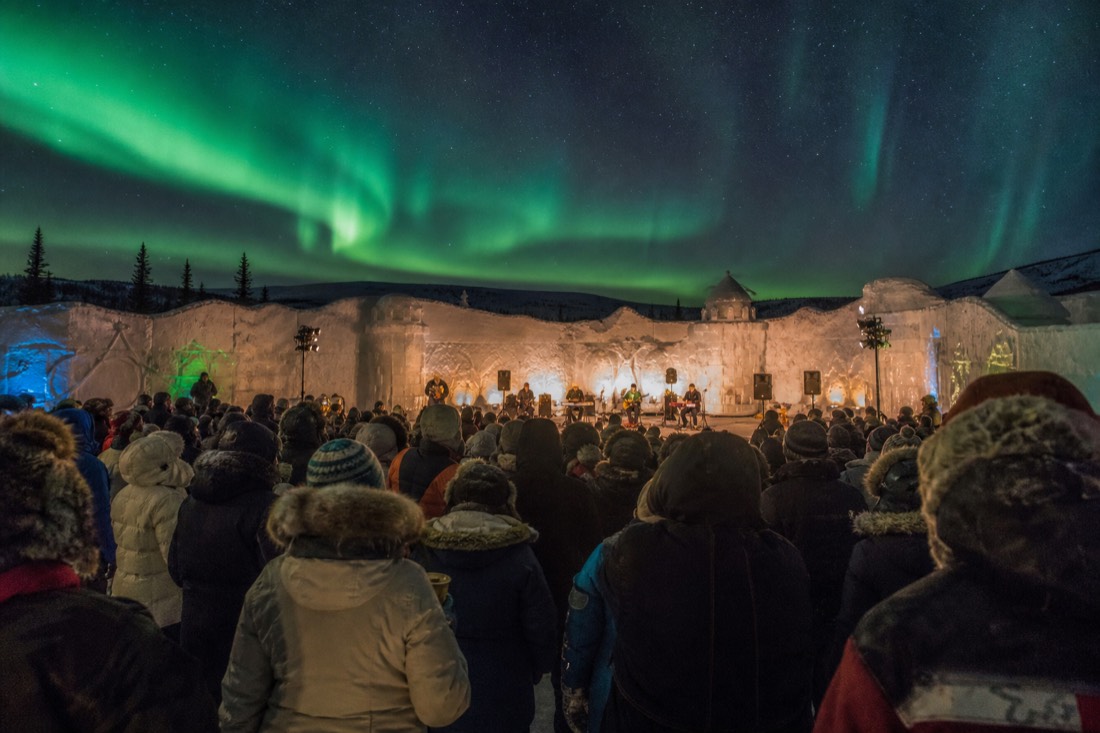 Concert crowd in parkas inside the snow castle courtyard with carved snow walls glowing in evening light