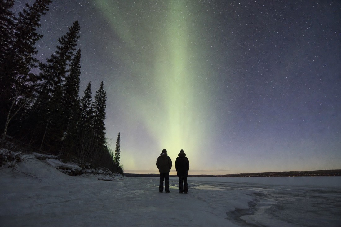 Two travelers silhouetted on a frozen lakeshore watching a towering pale green aurora column