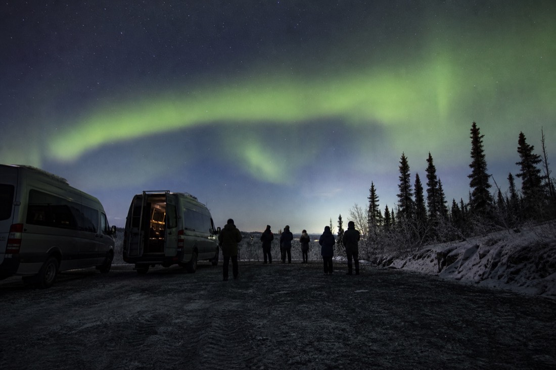 Two tour vans parked at a dark roadside pullout outside Yellowknife with guests watching a faint aurora arc overhead