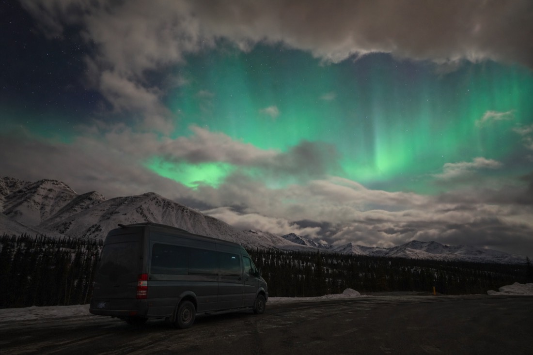 Tour van parked on a dark highway pullout under a breaking cloud sky with green aurora showing through the gap