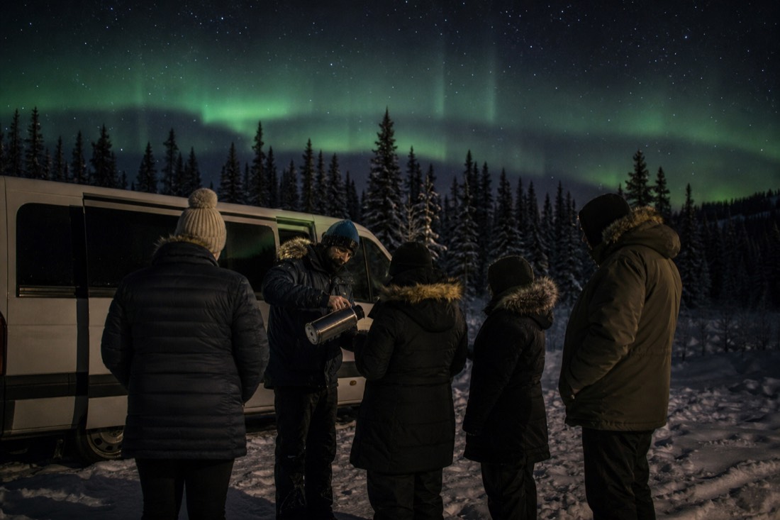 Guide pouring hot drinks from a thermos for guests beside a tour van while faint aurora builds overhead
