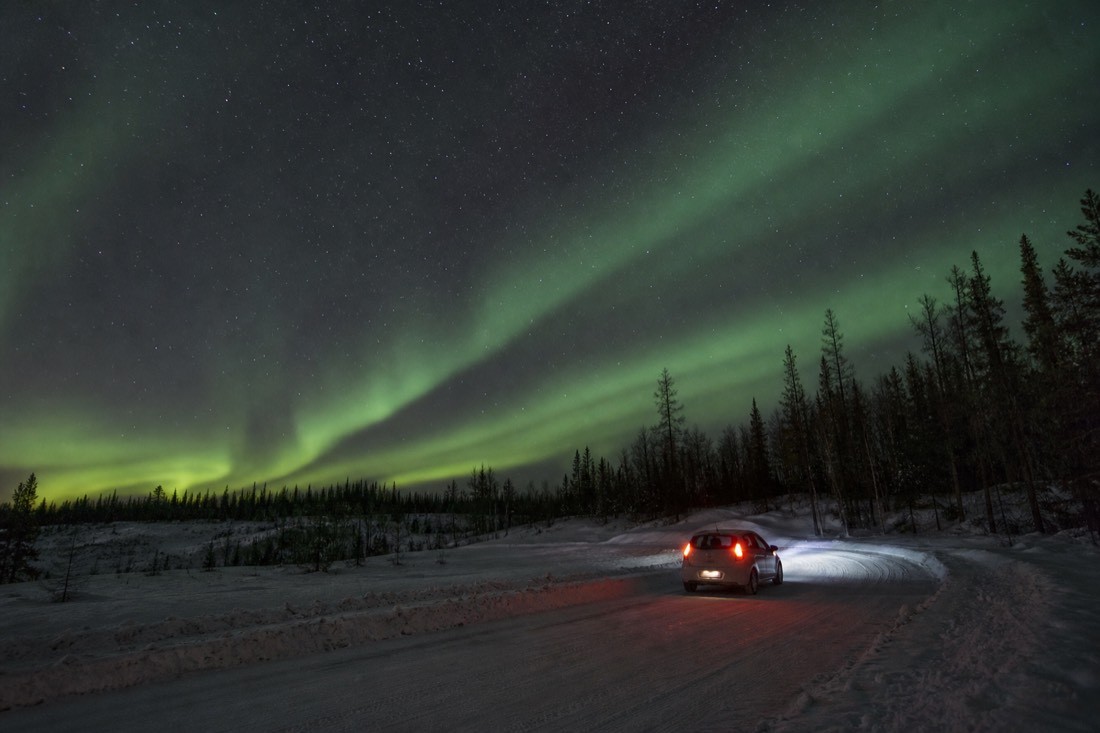 A small rental car driving a snow-packed road out of Yellowknife toward the Ingraham Trail in low winter sun