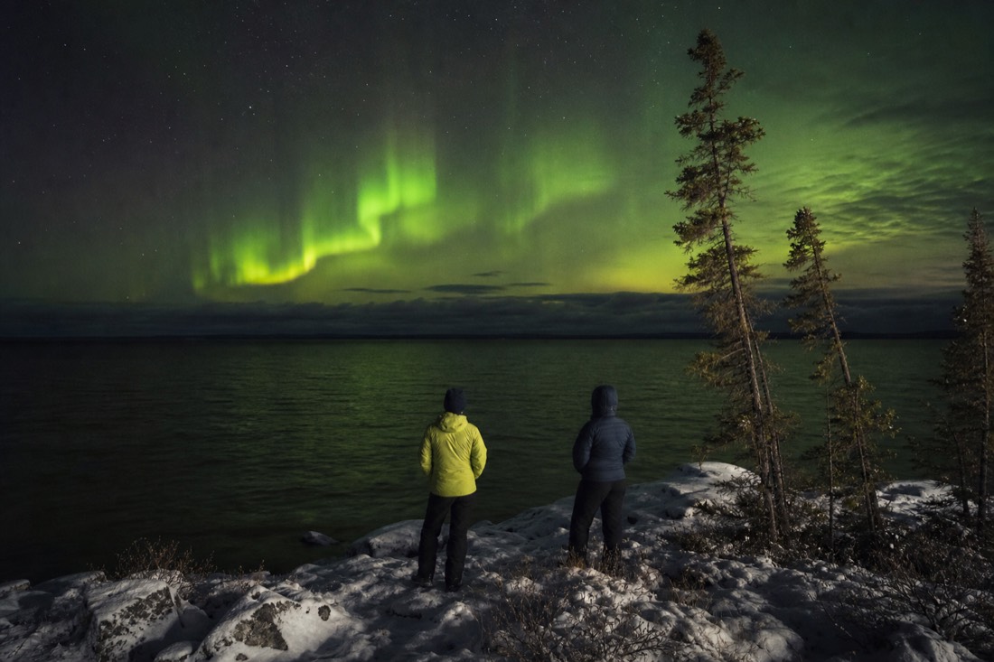 Autumn aurora over open water on Great Slave Lake with travelers in light jackets on the shore