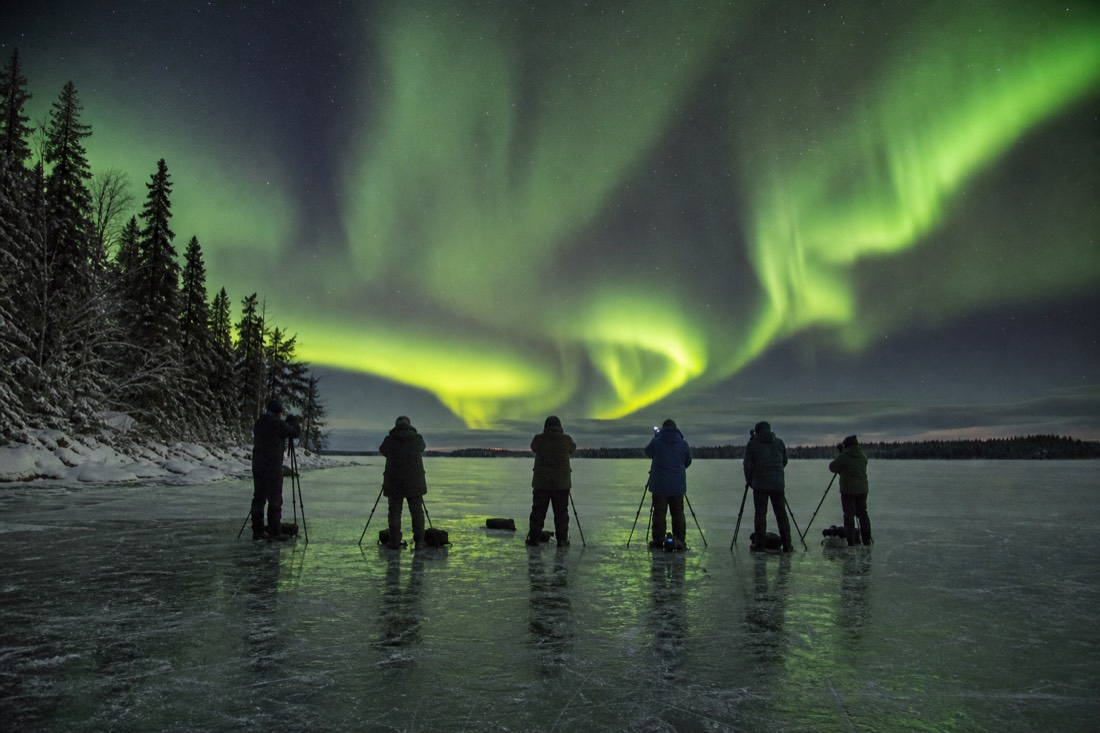 Row of aurora watchers with tripods on a frozen lake under a swirling green sky