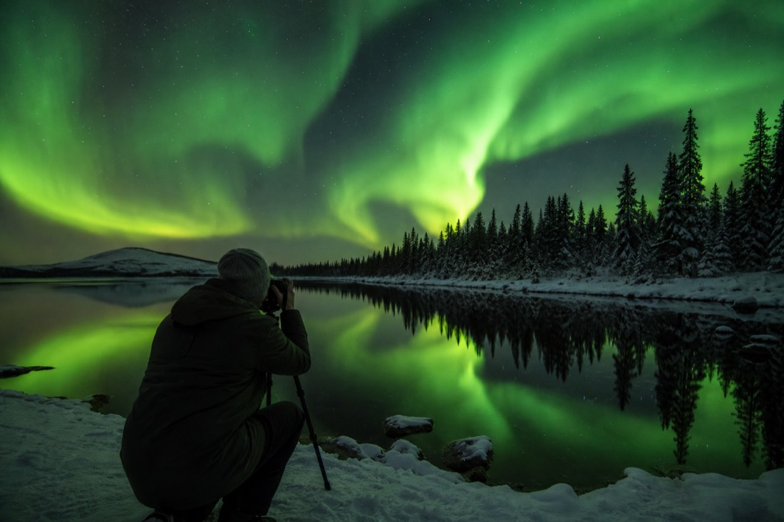 Traveler in a light jacket photographing aurora reflections on the still surface of a northern lake
